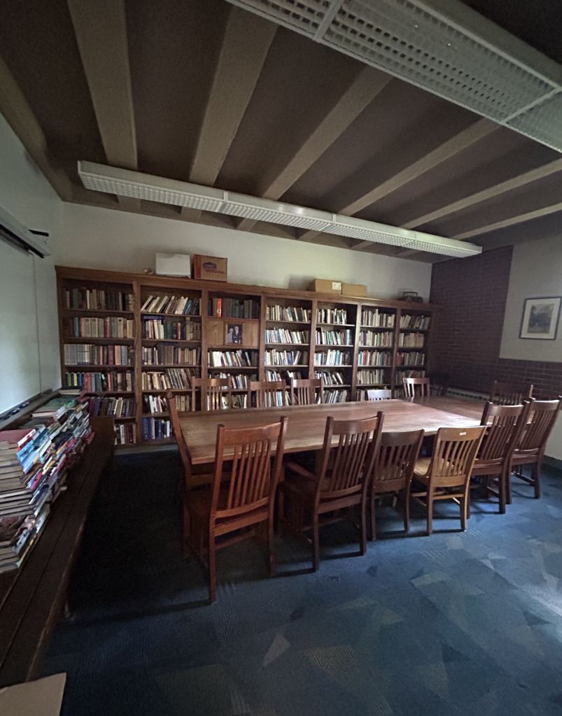 Sunlight streams onto a long, dark brown wooden table with dark brown wooden chairs on either side. A bookshelf stacked with books sits behind the table. 